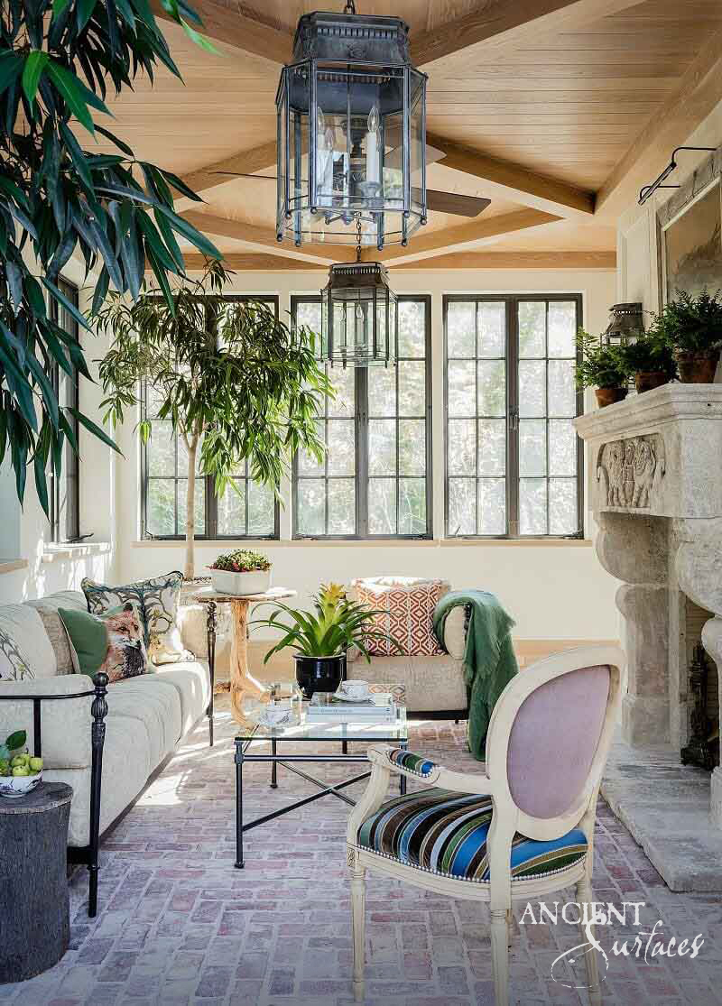 Reclaimed antique limestone mantel from Ancient Surfaces with a carved scrolled corbel in a sunlit garden room with a brick herringbone floor and hanging lanterns