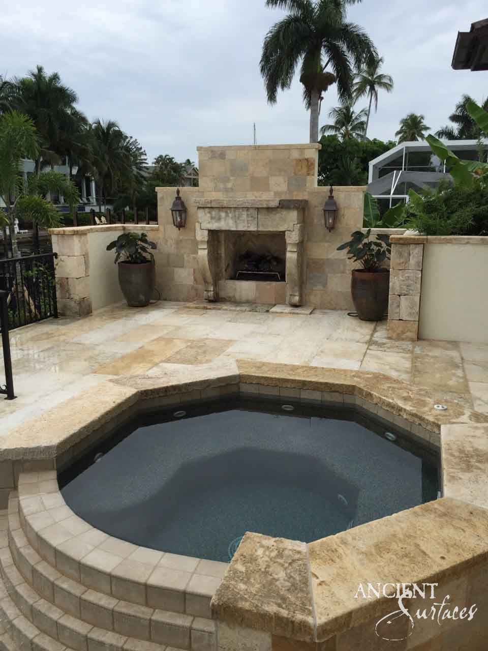 Reclaimed antique limestone fireplace from Ancient Surfaces built into a stacked stone surround beside an octagonal spa on a palm lined terrace