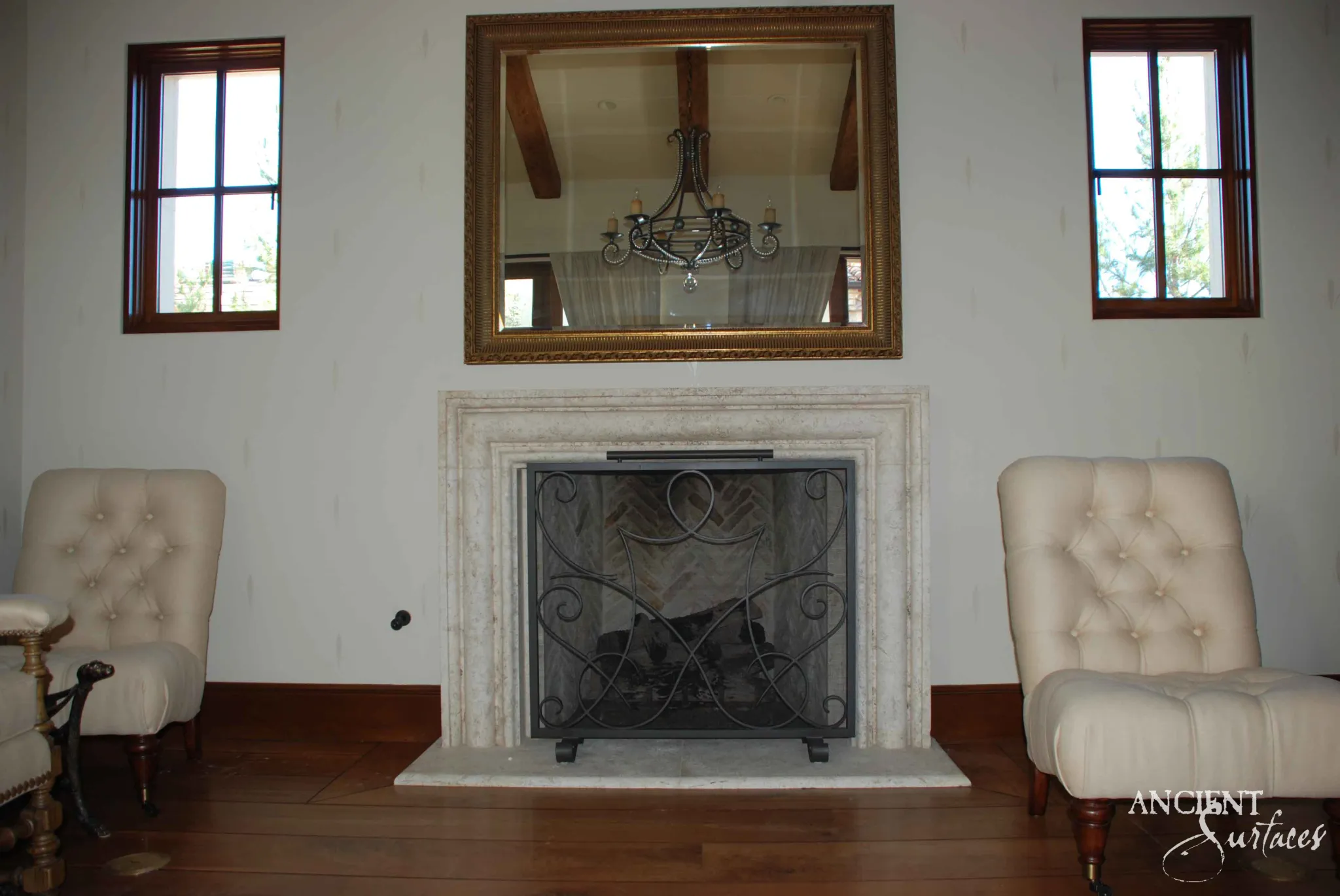 Reclaimed antique limestone mantel from Ancient Surfaces with a plain moulded surround beneath a gilt mirror flanked by cream tufted slipper chairs