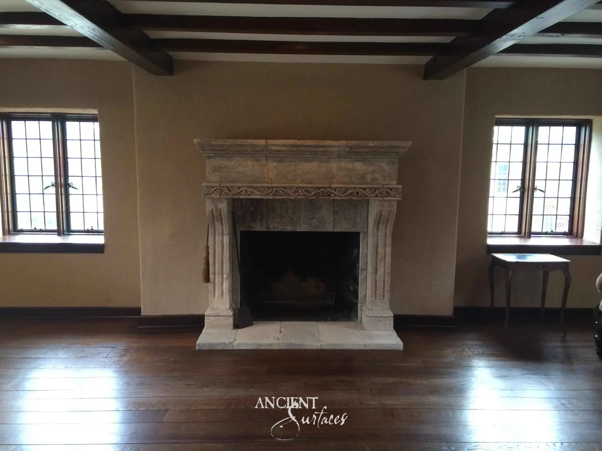 Reclaimed antique limestone mantel from Ancient Surfaces with a plain rectangular opening in an empty sunlit room with dark wood ceiling beams and leaded windows