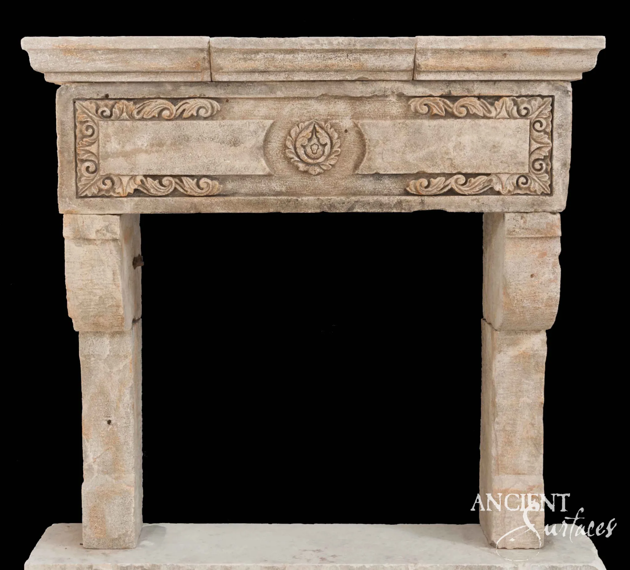 Reclaimed antique limestone mantel from Ancient Surfaces photographed against black with a carved wreath medallion and scrolling foliate corners on angled legs