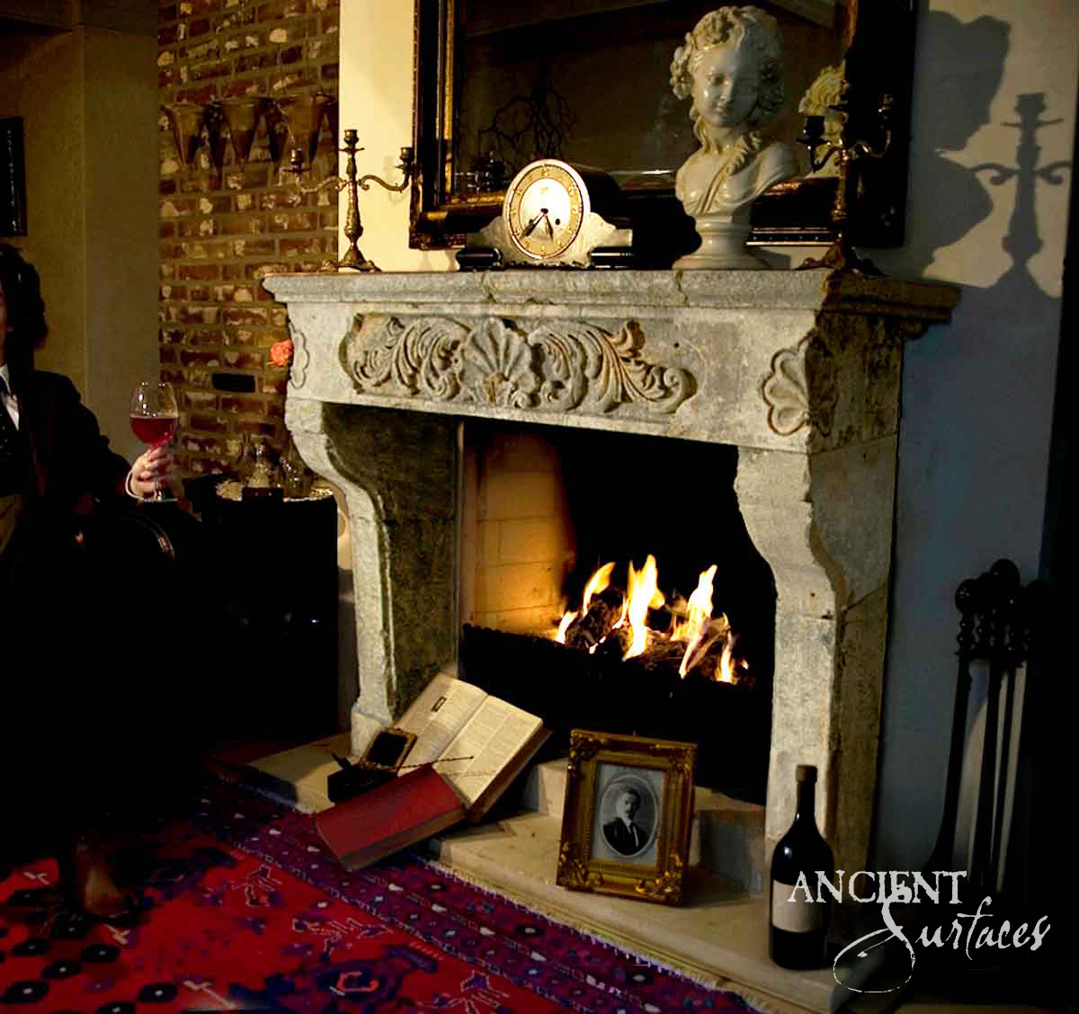 Reclaimed antique limestone mantel from Ancient Surfaces with a carved shell and scroll cartouche beside a lit fire in a brick chimney breast study