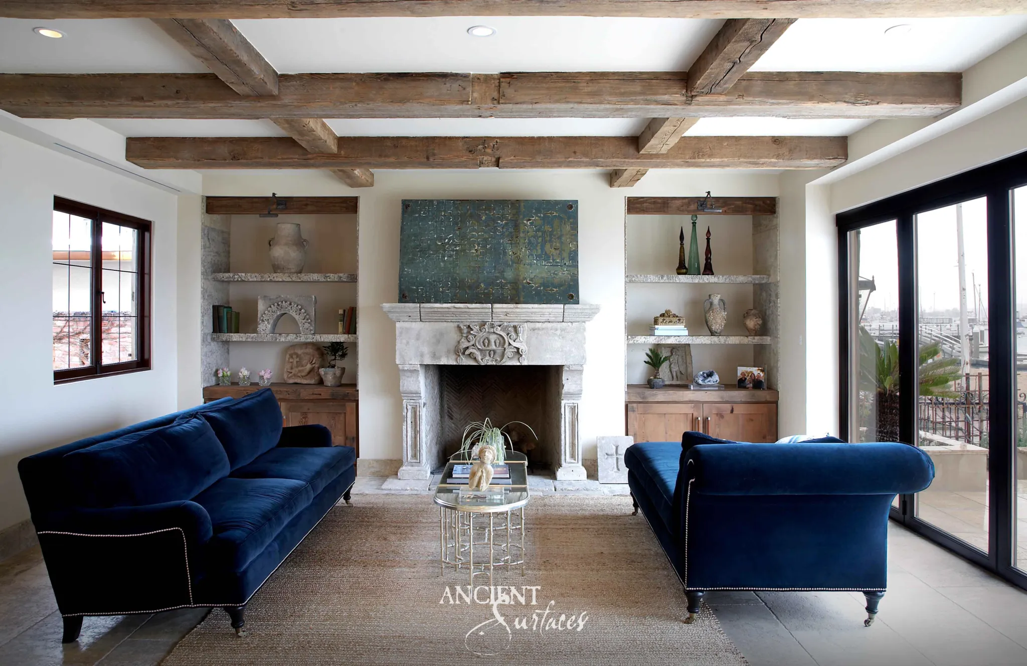 Reclaimed antique limestone mantel from Ancient Surfaces with a carved rosette medallion between blue velvet sofas in a living room overlooking a marina